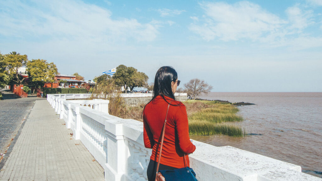 Mulher de blusa vermelha encostada em um parapeito branco observando o Rio da Prata, com vegetação à beira da água e uma bandeira do Uruguai ao fundo, no Centro Histórico de Colonia del Sacramento. Um local tranquilo e com pousadas charmosas, ideal para quem procura onde ficar em Colonia del Sacramento com comodidade.