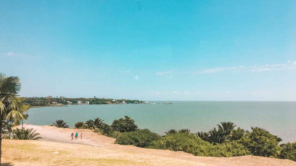 Vista panorâmica da costa e do Rio da Prata, na Rambla de Las Américas, com vegetação verde, céu azul e algumas pessoas caminhando por uma trilha de terra.