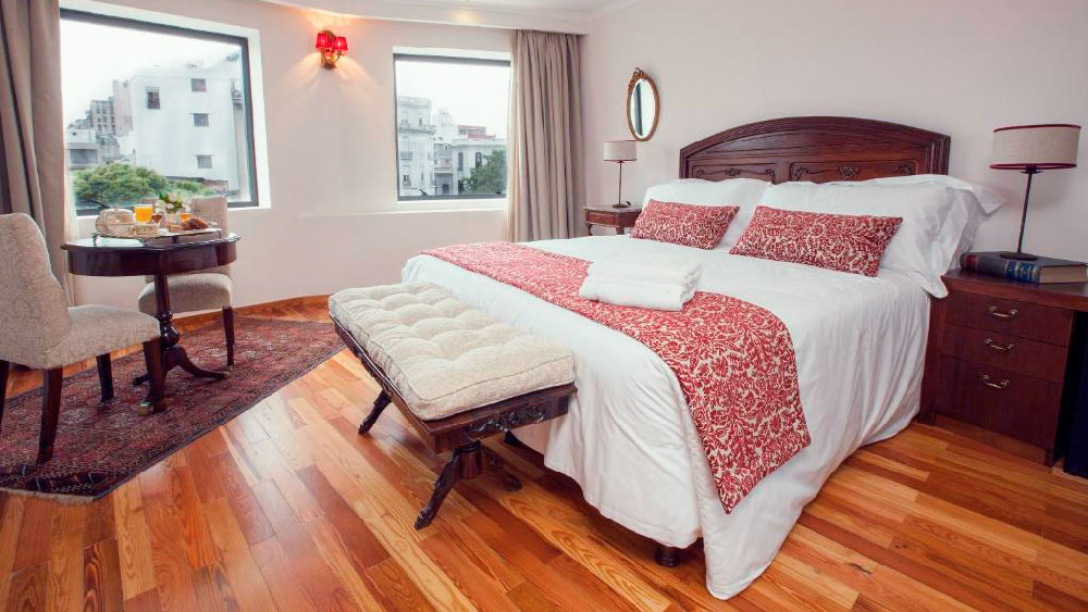 Quarto charmoso do Alma Histórica Boutique Hotel, um hotéis de Montevidéu mais charmoso, com cama de casal decorada com colcha e almofadas vermelhas estampadas, móveis de madeira escura e piso de madeira. Um canto com mesa de café da manhã e janelas amplas oferece vista para a arquitetura clássica do bairro.