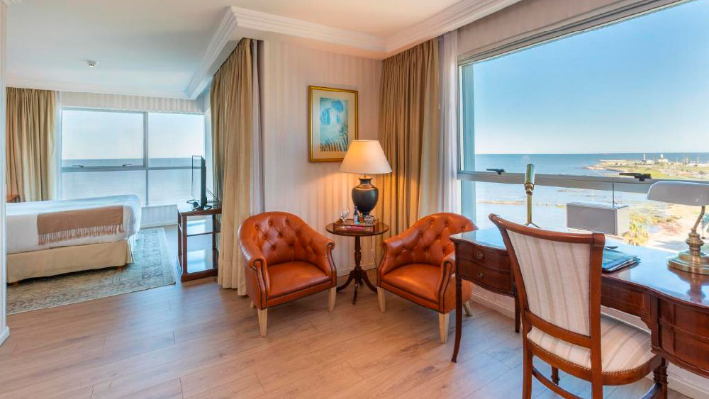 Suíte clássica do Cala di Volpe Boutique Hotel com vista ampla para o mar, mobiliada com poltronas de couro caramelo, escrivaninha de madeira e cama confortável ao fundo. As janelas grandes proporcionam luz natural e um panorama relaxante da costa de Montevidéu.