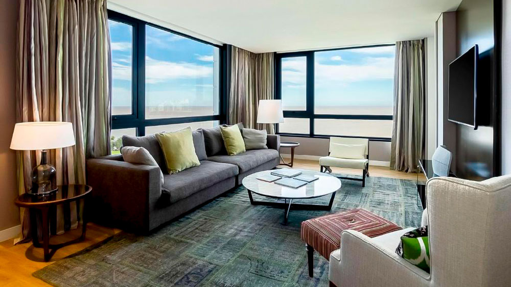 Sala de estar espaçosa da suíte no Hyatt Centric Montevideo, com sofá cinza, poltronas modernas e janelas amplas com vista para o Rio da Prata. A decoração mescla tons neutros com toques de cor e mobiliário contemporâneo, criando um ambiente confortável e iluminado.