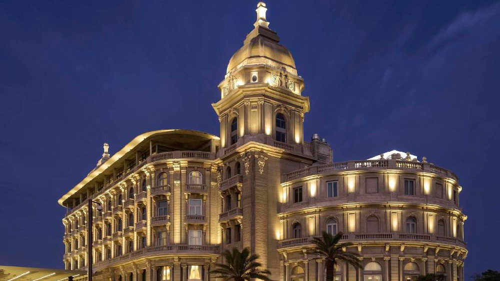 Fachada iluminada à noite do Sofitel Montevideo Casino Carrasco & Spa, com sua arquitetura imponente de estilo clássico europeu, cúpula central e detalhes ornamentados. O prédio histórico se destaca com palmeiras ao redor e céu azul profundo ao fundo, transmitindo elegância e prestígio.