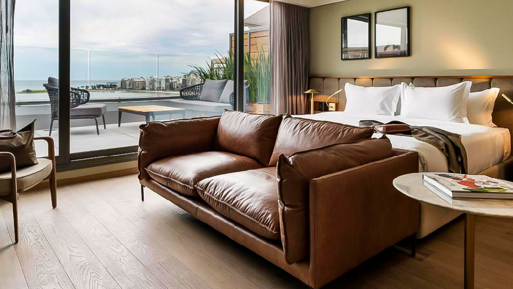 Suíte elegante do Costanero Hotel, um dos melhores hotéis de Montevidéu, com cama king-size, sofá de couro marrom e varanda espaçosa com vista para o mar e a cidade. A decoração combina tons neutros, móveis modernos e iluminação suave para um ambiente acolhedor e sofisticado.