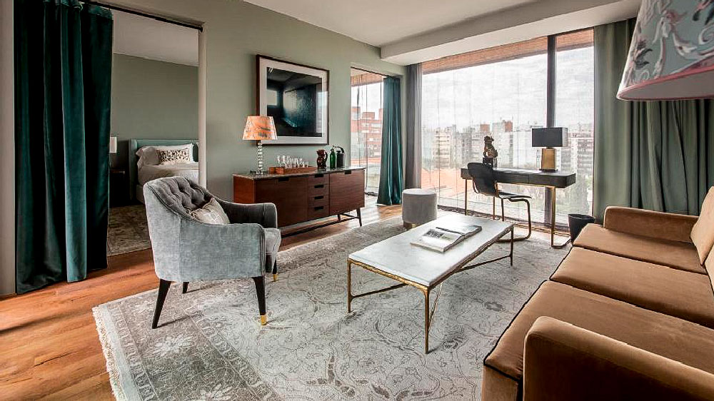 Sala de estar sofisticada da suíte do Hotel Montevideo, com decoração elegante e vista panorâmica da cidade através de janelas de vidro do chão ao teto. O espaço conta com sofá caramelo, poltrona cinza, mesa de centro com tampo claro, escrivaninha com luminária e quarto visível ao fundo.