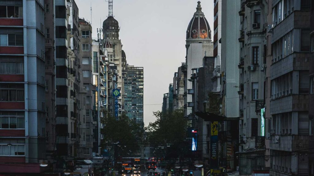 Vista de Montevidéu com prédios altos alinhados em ambos os lados da avenida, com destaque para duas torres antigas com cúpulas. A iluminação baixa do fim do dia cria um contraste entre os prédios.