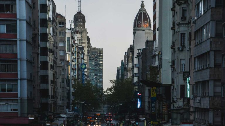 Vista de Montevidéu com prédios altos alinhados em ambos os lados da avenida, com destaque para duas torres antigas com cúpulas. A iluminação baixa do fim do dia cria um contraste entre os prédios.